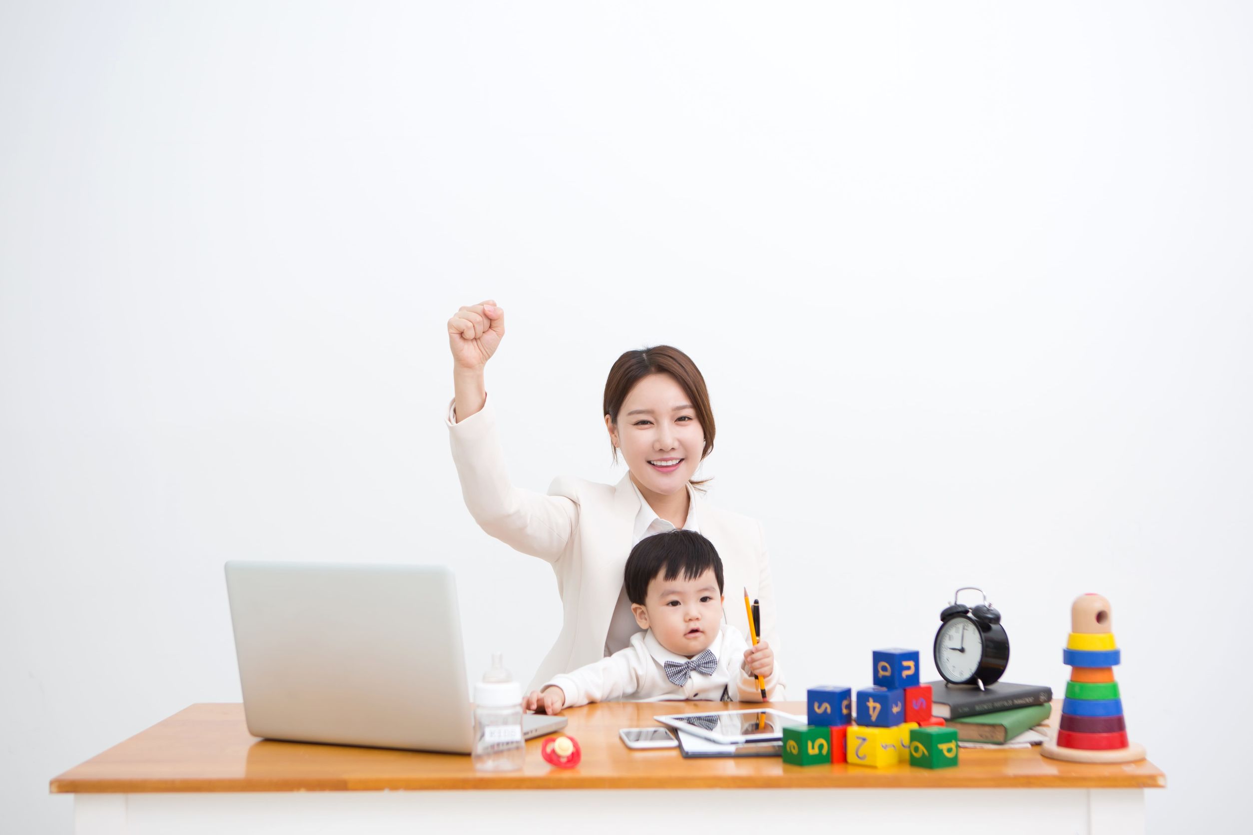 一位年輕母親抱著兒子在辦公桌前握拳微笑，桌上擺放著筆電、嬰兒玩具、鐘和書本，象徵工作與育兒平衡。